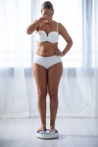 Woman in white underwear covering her eyes on a weighing scale